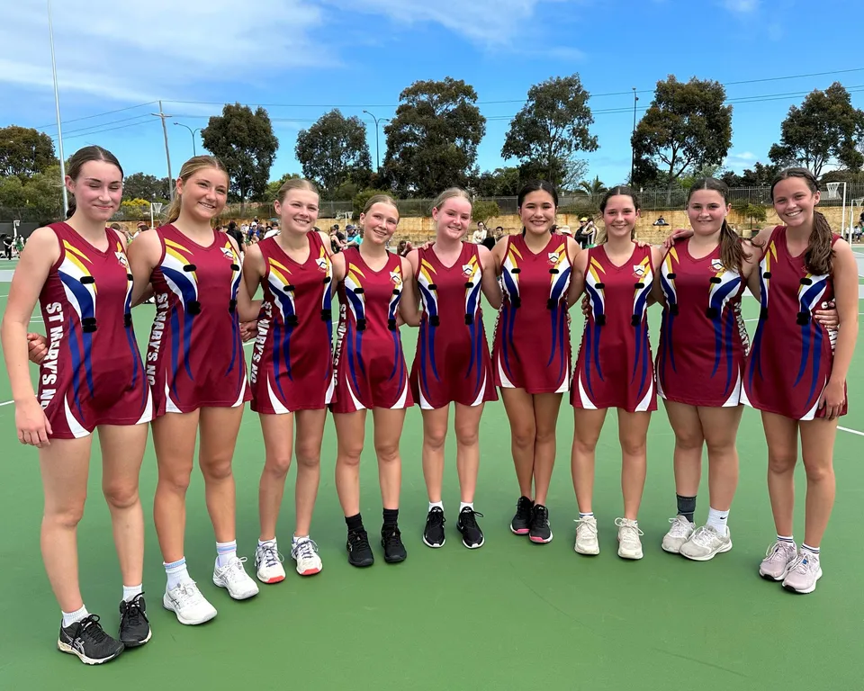 Un gruppo di giovani atlete in maglia rossa e blu si trova insieme sul campo sportivo della St. Mary's Anglican Girls' School.