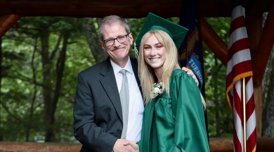 Un uomo anziano in giacca e cravatta è accanto a una giovane donna con abito da laurea verde nel campus della Leelanau School.