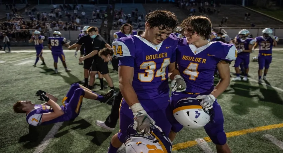 Due giocatori di football si trovano sul campo da gioco delle Boulder Valley Public Schools con giocatori e spettatori sullo sfondo.