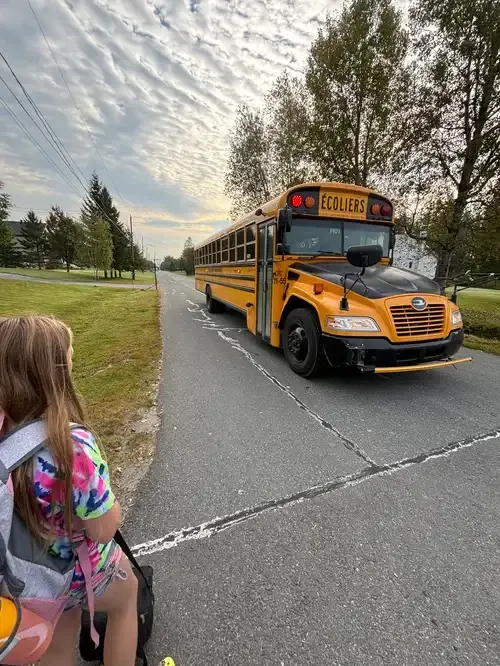 Chica con mochila esperando junto a la carretera a un autobús escolar amarillo