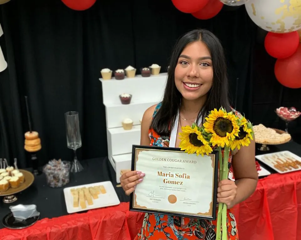 Una studentessa della John F. Kennedy High School tiene in mano un certificato e dei girasoli davanti a un tavolo con dolci e decorazioni.