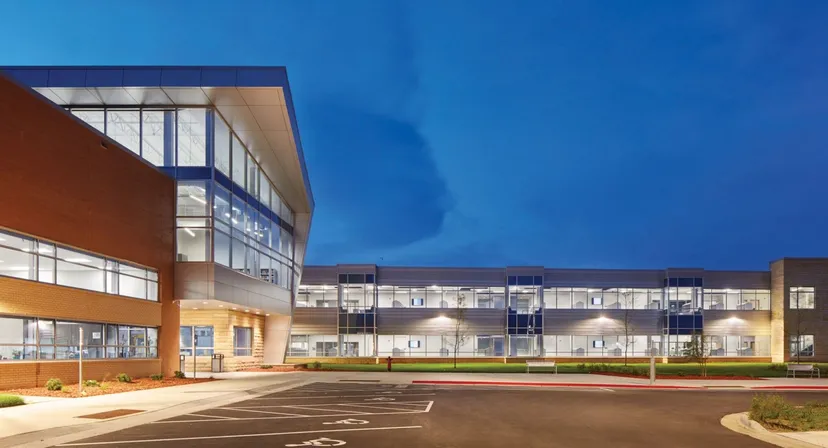 L'edificio moderno a più piani della St. Peter High School, con ampie vetrate e facciata arancione, si staglia sotto un cielo azzurro e limpido.