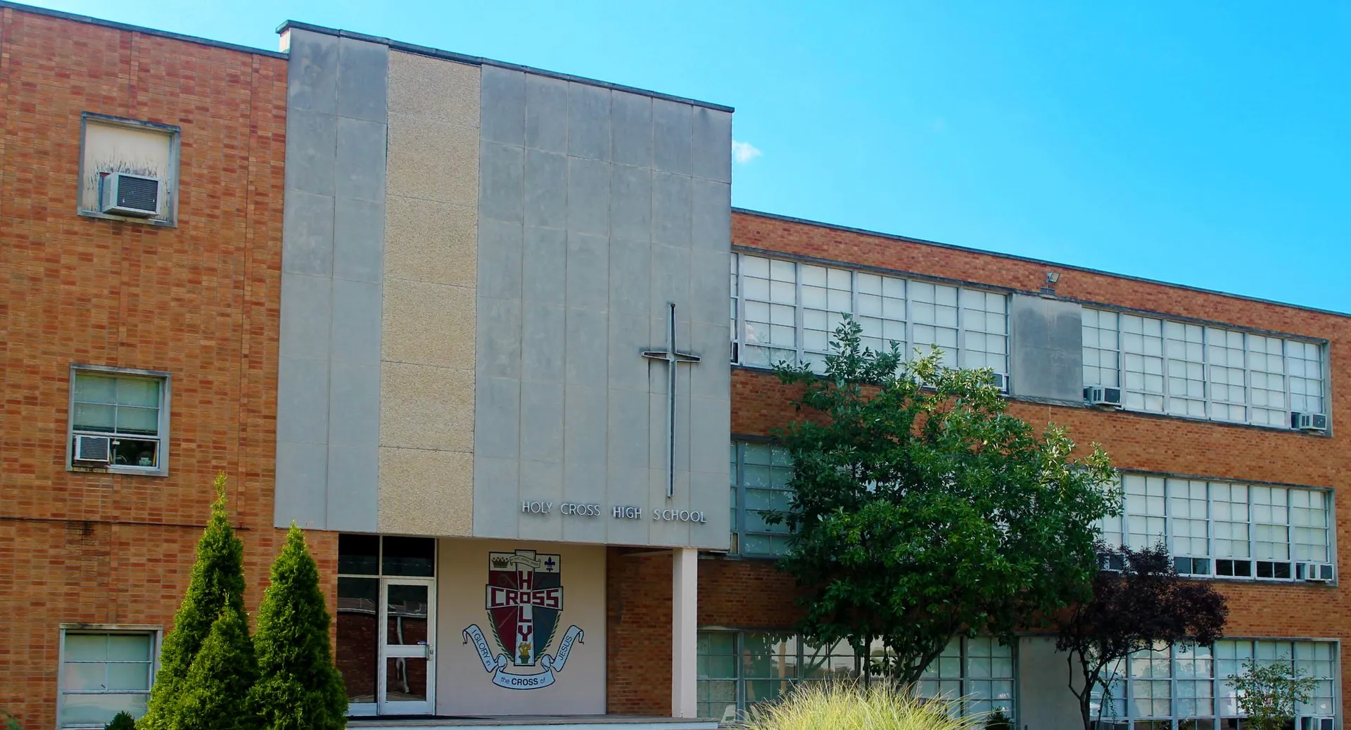 L'edificio in mattoni a più piani della Holy Cross High School, con grandi finestre e un ingresso caratteristico, sorge circondato da alberi sotto un cielo limpido.