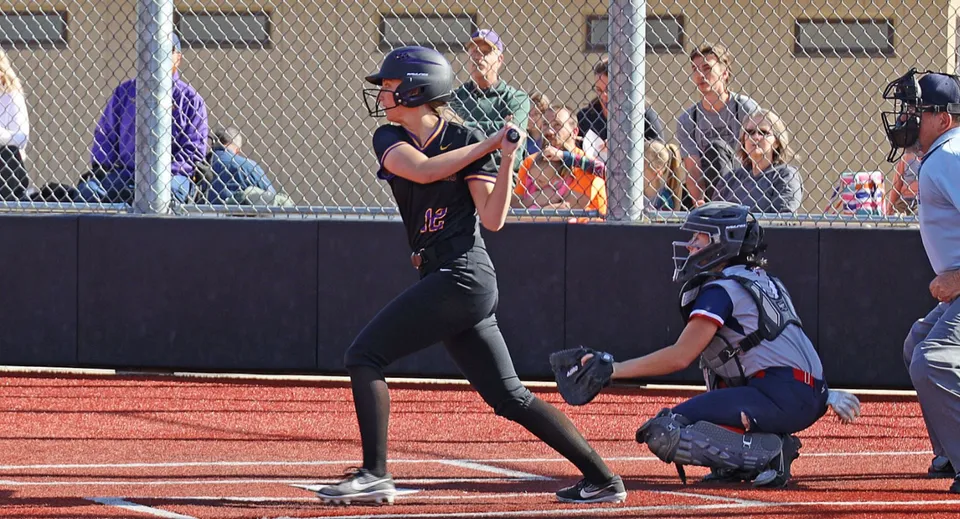 Un giocatore di softball con la divisa nera si trova sul campo della Cascade Christian High School con un ricevitore in divisa blu dietro la casa base.