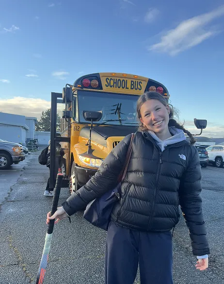 Chica sonriendo con un palo de hockey delante de un autobús escolar amarillo