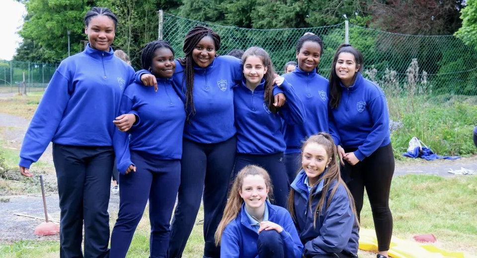 Un gruppo di studentesse della Our Lady's Bower Secondary School si trova all'aperto su un prato con alberi e una recinzione sullo sfondo.