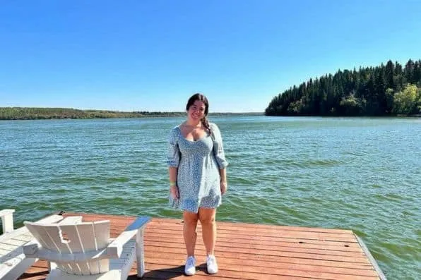 Joven sonriente con vestido azul sobre un embarcadero de madera junto a un amplio lago