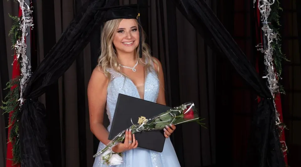 Una diplomata della W.R Myers High School è in piedi davanti a una tenda nera con un abito blu, il tocco da diploma, un diploma e una rosa rossa.