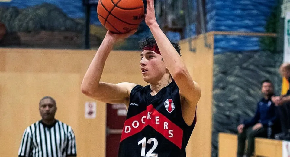 Un alunno della Esquimalt High School salta per tirare a canestro sul campo da basket con gli spettatori sullo sfondo.