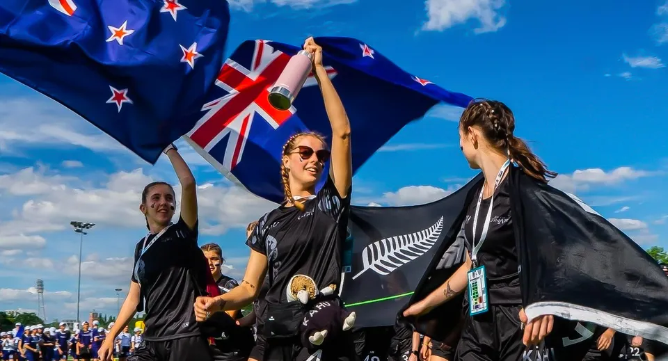 Un gruppo di studenti e studentesse del Waitakere College saluta sventolando la bandiera neozelandese sotto un cielo blu con la Union Jack e la bandiera con la felce d'argento.