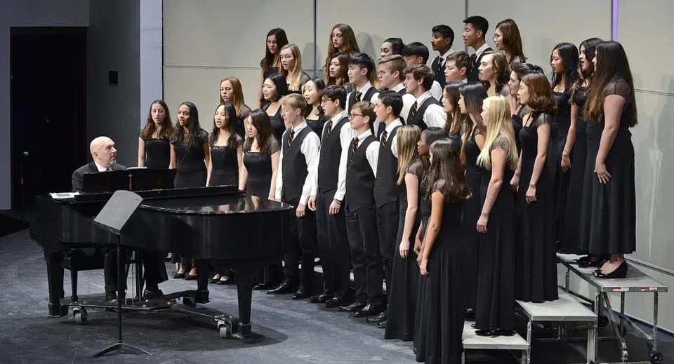 Un grande coro in abiti formali neri sta sul palco della Lake Forest Academy con un pianoforte a coda in primo piano.