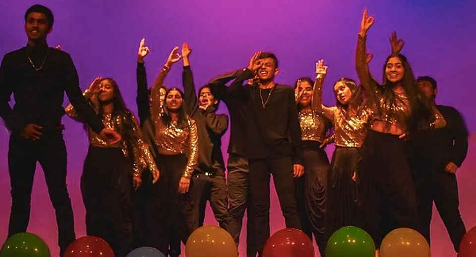 Studenti del Woburn Collegiate Institute sono sul palco con costumi nero-oro e le braccia alzate davanti a uno sfondo lilla.
