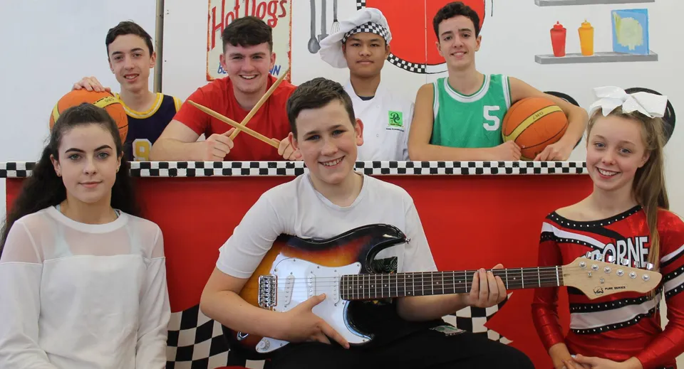 Un gruppo di studenti e studentesse con strumenti musicali e attrezzature sportive davanti a un motivo a scacchi colorato nel campus del Davitt College.