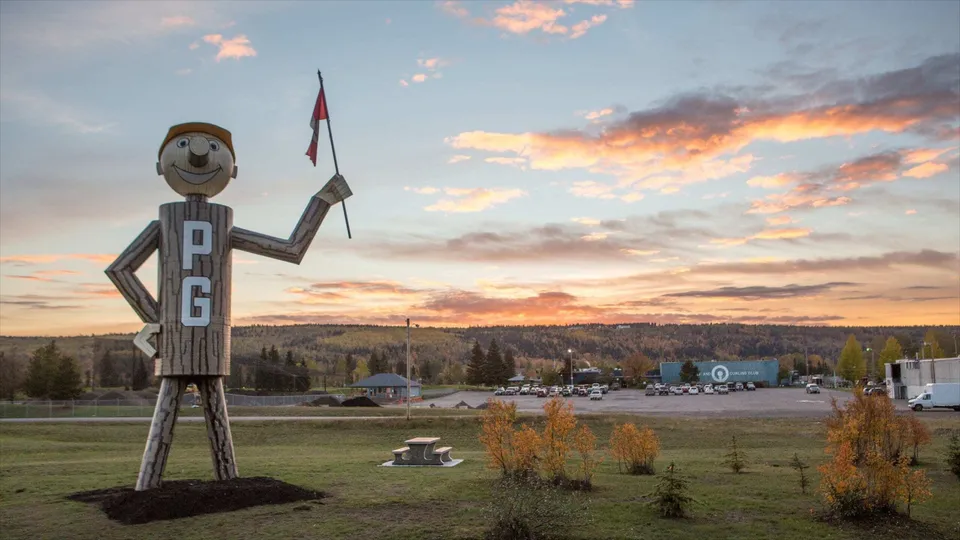 Una grande figura in legno con un viso sorridente si trova davanti a un paesaggio con montagne e alberi nel terreno della Duchess Park Secondary School.