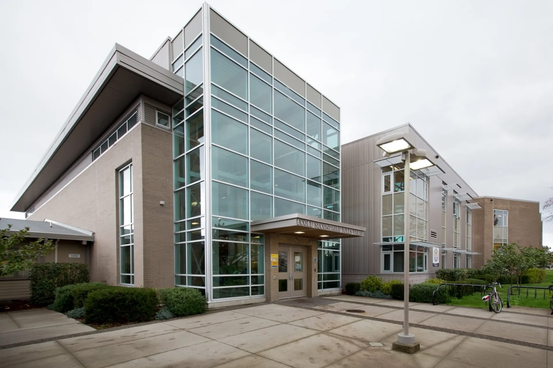 Il moderno edificio a più piani con facciata in vetro della Jules Verne School Vancouver è circondato da un vialetto pavimentato e aree verdi.