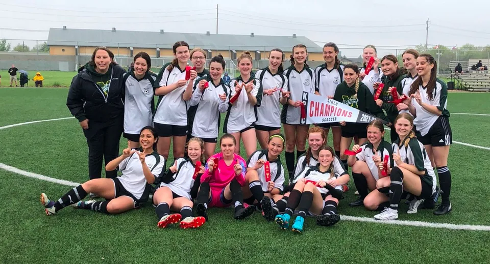 Un gruppo di giovani calciatrici del Miles Macdonell Collegiate festeggia la vittoria sul campo con uno striscione del campionato.