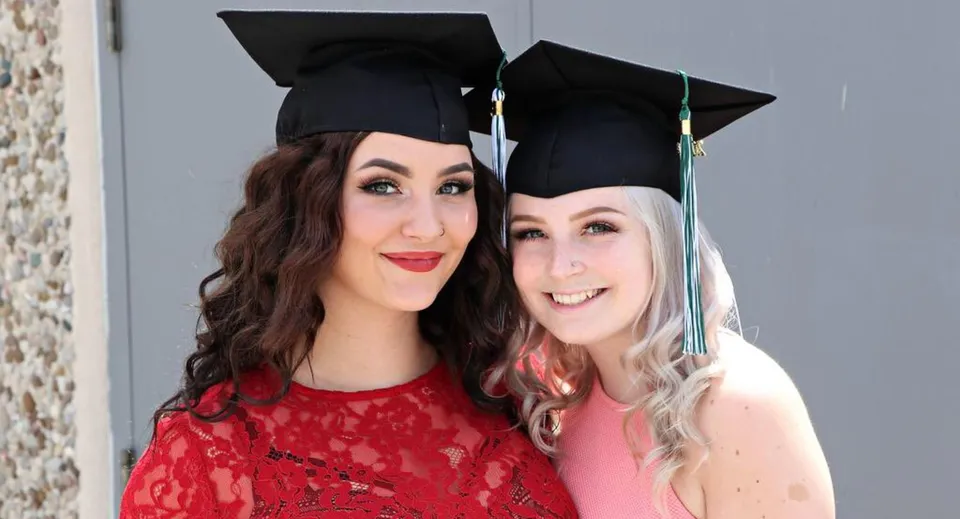 Due studentesse della W.F. Herman Academy Secondary School posano sorridenti con il tocco da diploma davanti a una parete grigia.