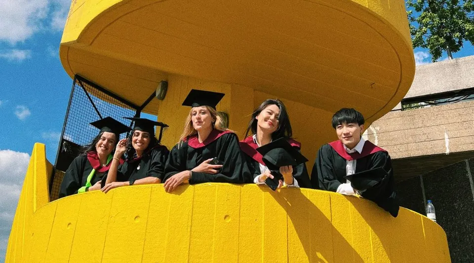Un gruppo di neolaureati e neolaureate della University of Westminster in toga rossa si trova davanti a un edificio giallo.
