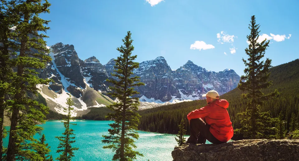 Una persona con una giacca rossa seduta su una roccia con vista su un lago turchese circondato da montagne innevate nel terreno della West Carleton Secondary School.