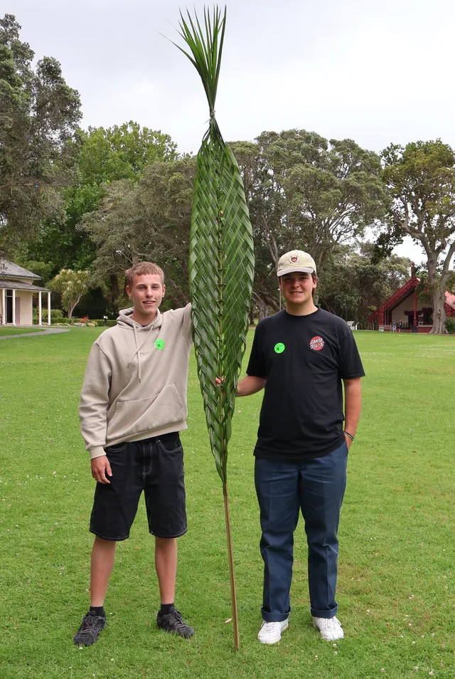 Dos chicos sujetando una gran hoja de lino trenzada en el césped ante una casa maorí
