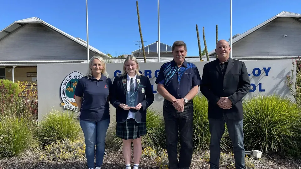 Una studentessa della Georgiana Molloy Anglican School posa con un premio insieme a tre adulti davanti al logo della scuola.