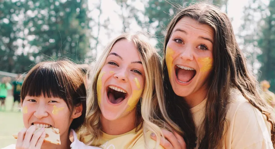Tre studentesse della Northwest Christian High School con pittura facciale gialla sorridono all'aperto.