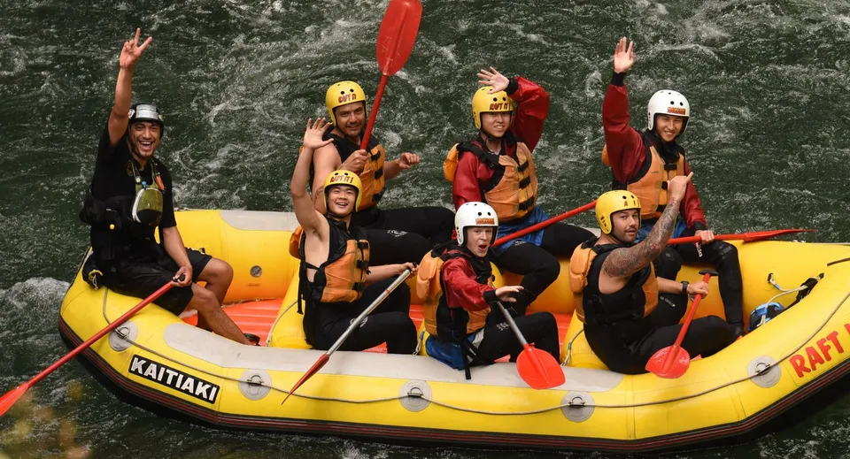 Studenti del Whangaparāoa College seduti con giubbotti salvagente e caschi in un gommone giallo in un paesaggio boschivo.