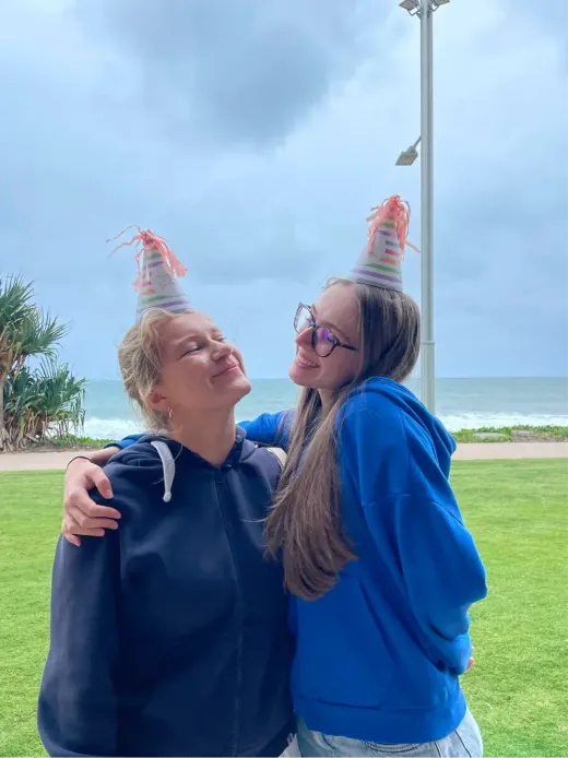 Zwei Mädchen mit Papierhütchen umarmen sich auf einer Wiese vor dem Strand in Australien