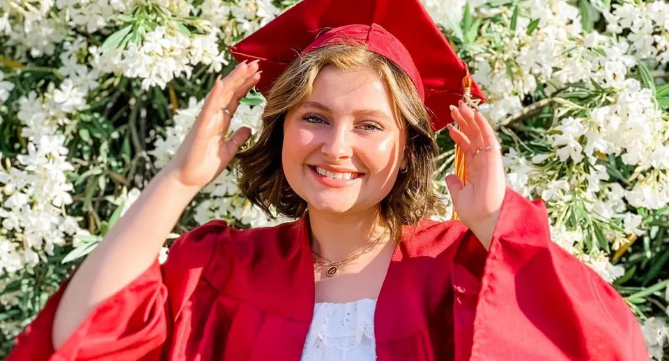 Una giovane donna sorridente in toga rossa da diploma davanti a fiori bianchi in fiore nel campus della Kings Christian School.