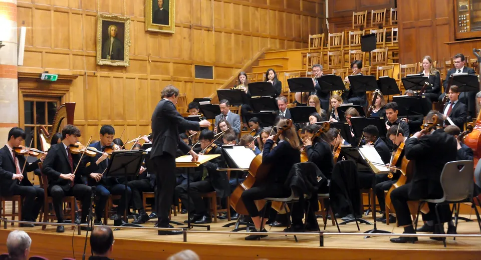 Un'orchestra studentesca della Rugby School suona sotto la direzione di un direttore in una sala rivestita di legno.