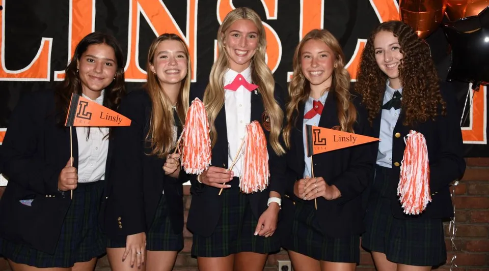 Un gruppo di studentesse della The Linsly School in divisa nera con dettagli rosa sta davanti a un grande cartello Lindy tenendo in mano pompon rosa.