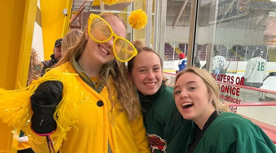 Tre studentesse della Rideau District High School posano sorridenti in un'arena sportiva con attrezzi sportivi sullo sfondo.