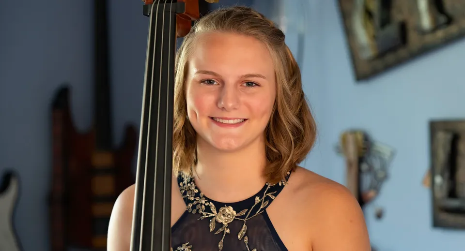 Una giovane donna tiene un violino davanti a una parete con opere d'arte incorniciate nel Queensbury Union Free School District.