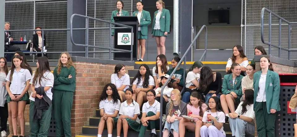 Studentesse della Asquith High School sedute su delle scalinate mentre tre compagne in divisa verde parlano da un podio.