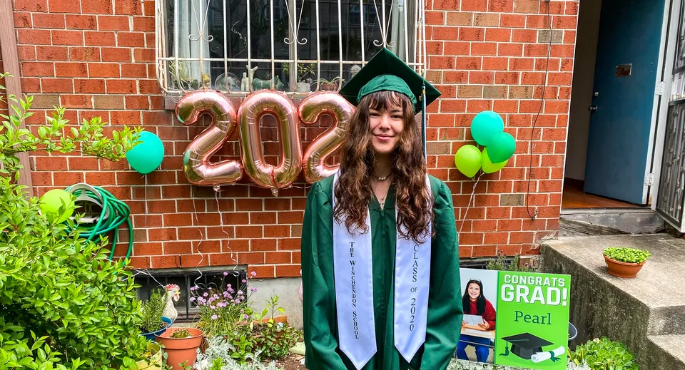 Una studentessa della Winchendon School a New York City indossa la toga verde da diploma davanti a un muro di mattoni con palloncini colorati e un cartello di congratulazioni.