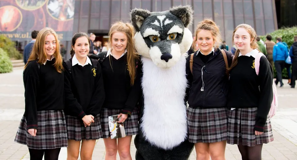 Studentesse della Scoil Aireagail in piedi insieme a un grande animale simile a un lupo bianco e grigio in primo piano.