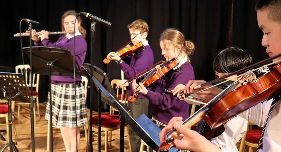 Un gruppo di musicisti in outfit viola suona sul palco dell'Ellesmere College diversi strumenti, come violini e batteria.