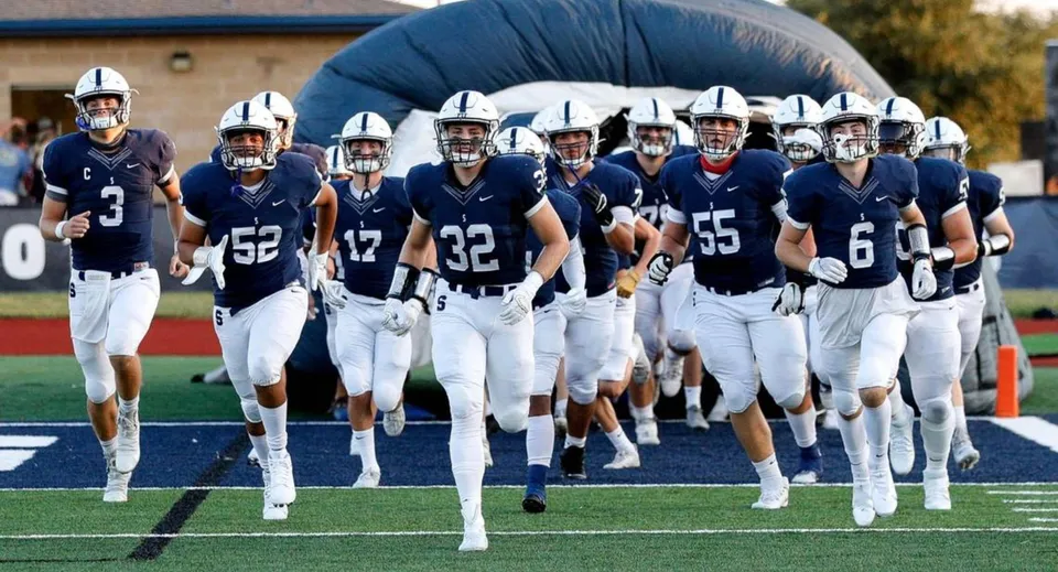 Un gruppo di giocatori di calcio della All Saints High School corre insieme su un campo da calcio con terreno di gioco blu e bianco.