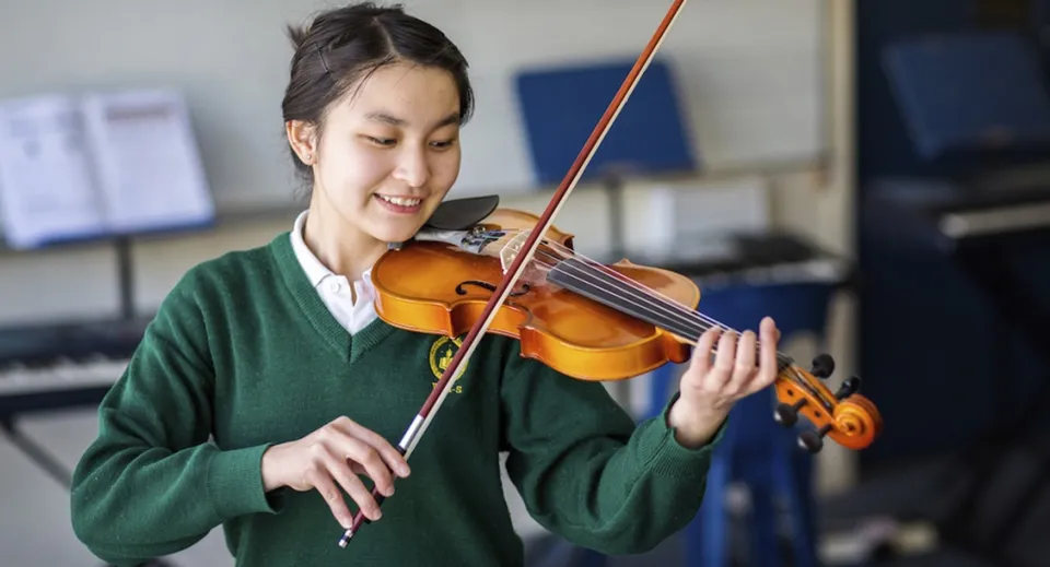 Una studentessa della Whanganui High School suona il violino davanti a strumenti musicali sullo sfondo.