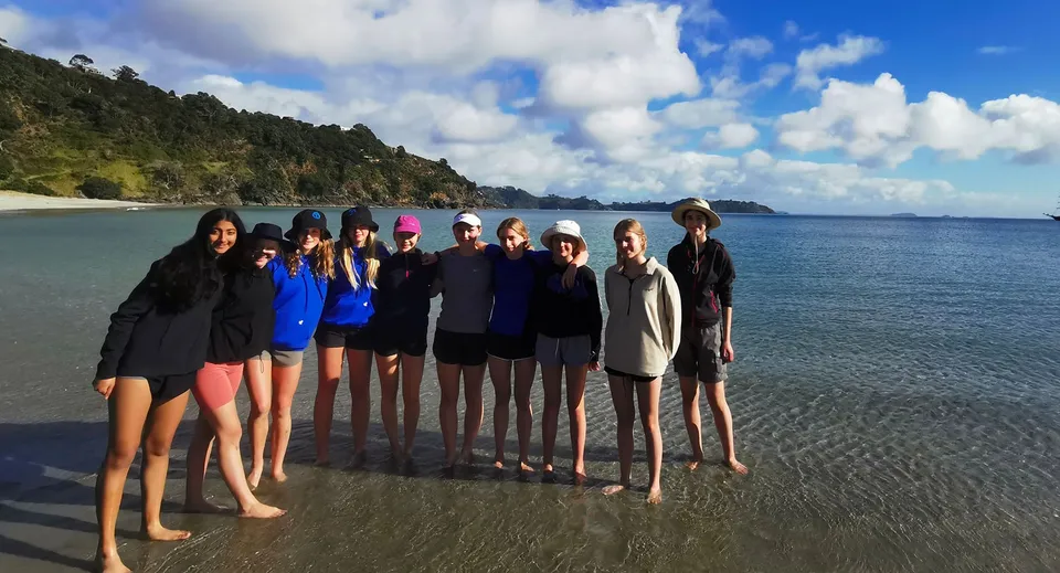 Un gruppo di studentesse del Wellington Girls' College si trova insieme sulla spiaggia con le montagne sullo sfondo.