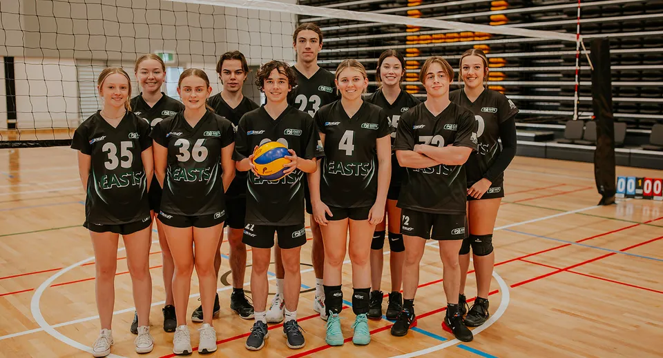 Un gruppo di giovani pallavoliste del Reynella East College si trova insieme sul campo da pallavolo con rete e tabellone segnapunti sullo sfondo.