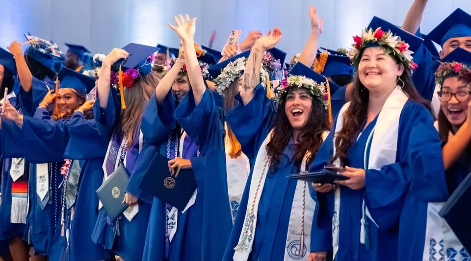 Un gruppo di neolaureati della Hawai'i Pacific University festeggia con le braccia alzate, indossando toghe blu e collane di fiori.