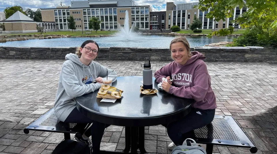 Due studentesse della SUNY Plattsburgh sono sedute a un tavolo davanti a una fontana e mangiano uno snack nel campus.