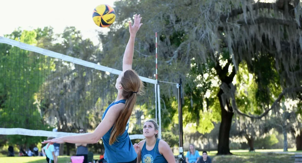 Una studentessa della Trinity Catholic High School colpisce un pallone da pallavolo in un parco con alberi e un'area verde.