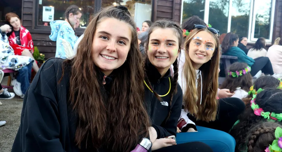 Tre studentesse con i capelli lunghi sorridono davanti a un edificio con facciata in legno sulla ringhiera della Presentation Secondary School a Kilkenny.
