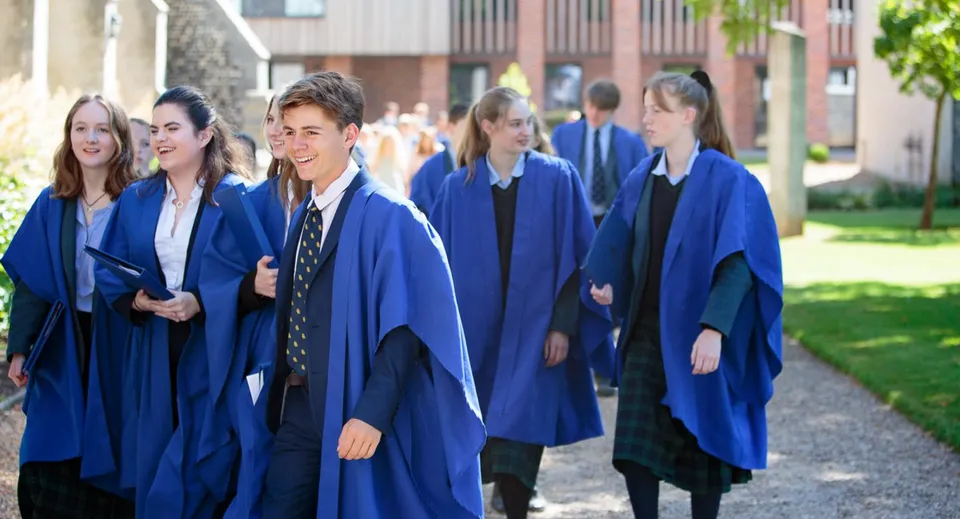 Un gruppo di studenti della Gresham’s School in toghe blu cammina su un viale tra edifici e aree verdi.