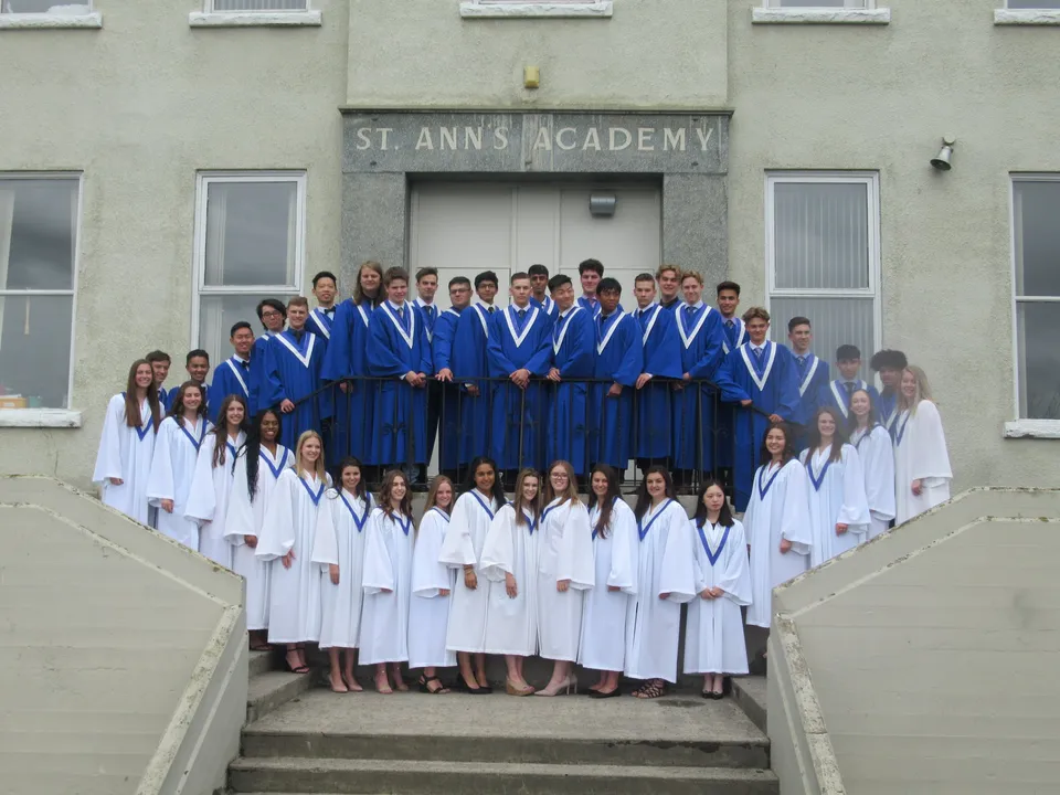 Un gruppo di studenti con toghe di laurea blu e bianche posa sulla scalinata della St. Ann's Academy.
