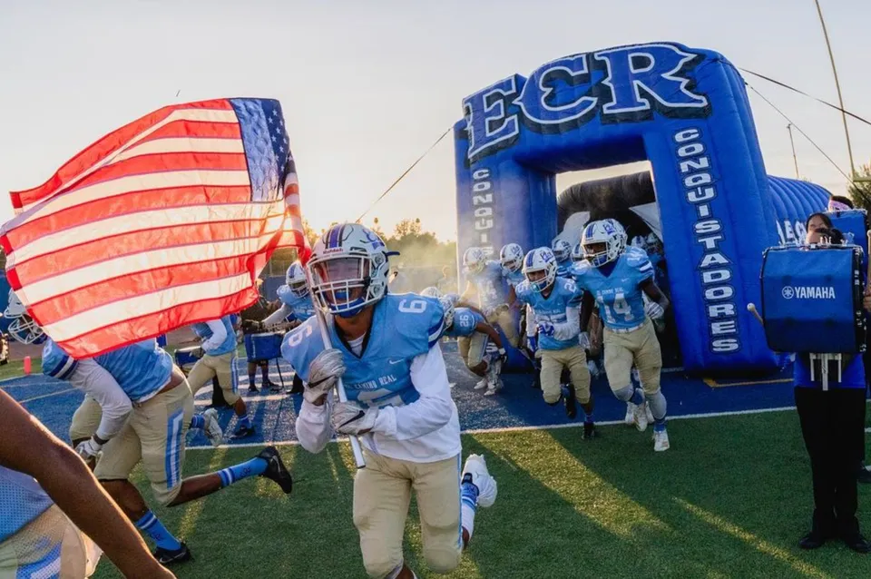 La squadra di calcio della El Camino Real Charter High School corre attraverso un tunnel gonfiabile con la bandiera americana in primo piano.