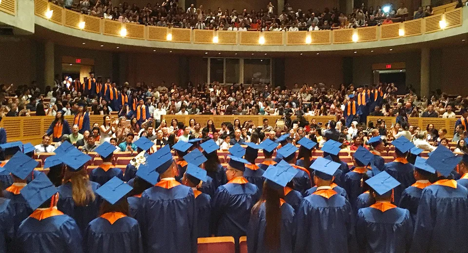 Diplomati e diplomate della Windermere Secondary School siedono in un'aula magna decorata a festa indossando la toga di diploma blu e arancione.