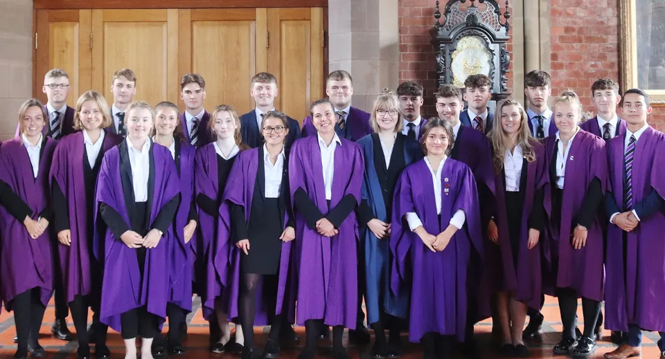 Un gruppo di diplomati in toga viola sta davanti a un edificio in mattoni a vista con un arco decorato nel campus dell'Ellesmere College.